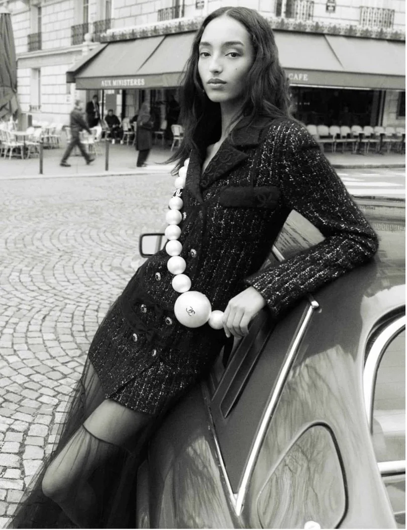 Black-and-white photograph of a model leaning on a vintage car in Paris, from Chanel Fall-Winter 2025/26, photographed by Mikael Jansson.