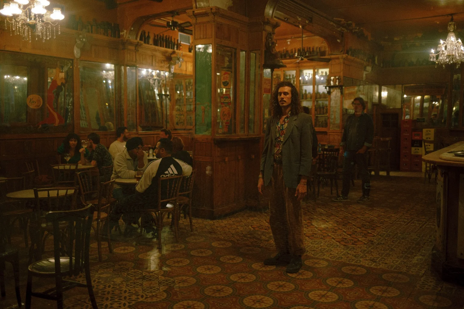 A warm, cinematic photograph of Mateo Aimaretti standing in the middle of a vintage bar, surrounded by people talking at small tables under chandeliers. Photo by Marina Klcheva.