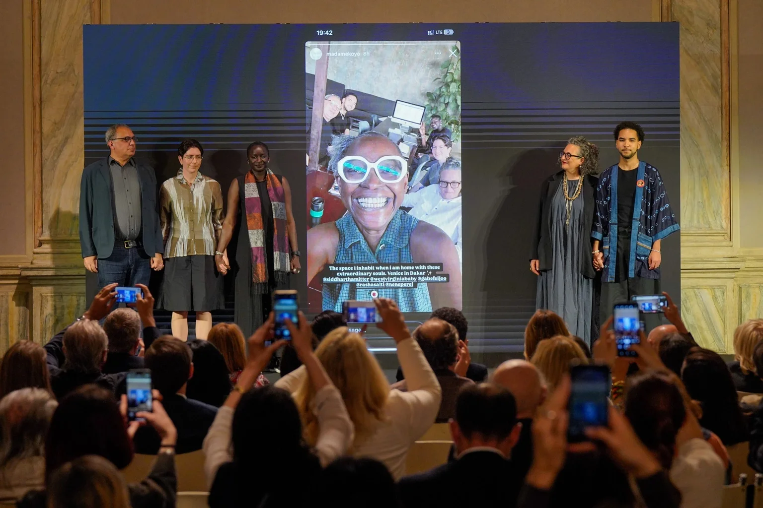 The curatorial team of Biennale Arte 2026 standing on stage in front of a large screen, while an audience photographs them with their phones. Photo by Andrea Avezz, courtesy of La Biennale di Venezia.