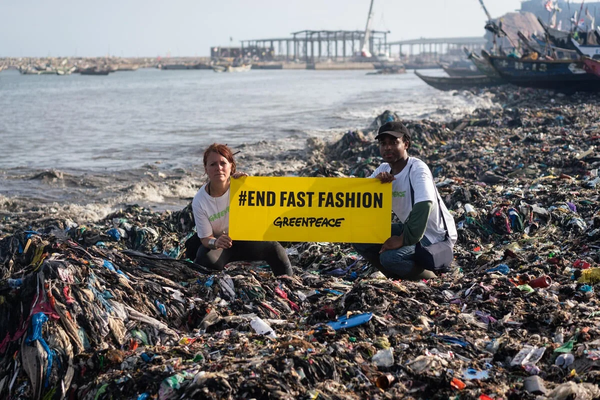 Two Greenpeace activists kneeling on a beach covered in textile and plastic waste, holding a yellow banner that reads #END FAST FASHION Greenpeace.