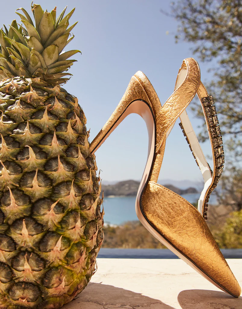 Close-up of gold high heels leaning against a pineapple with the sea and hills in the background, illustrating pineapple-based Piatex as a sustainable material.