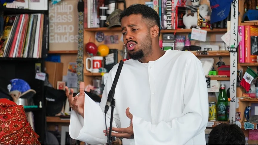 Mustafa performing at his NPR Tiny Desk Concert, singing into a microphone while wearing a white thobe, surrounded by shelves of books and objects.