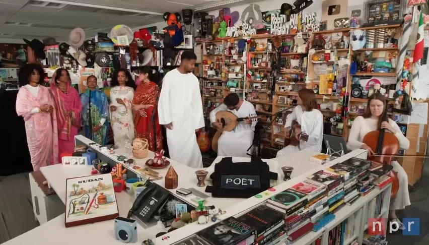 Wide view of Mustafas NPR Tiny Desk Concert, showing Mustafa in a white thobe with musicians and family members in colorful Sudanese garments gathered around the Tiny Desk set.