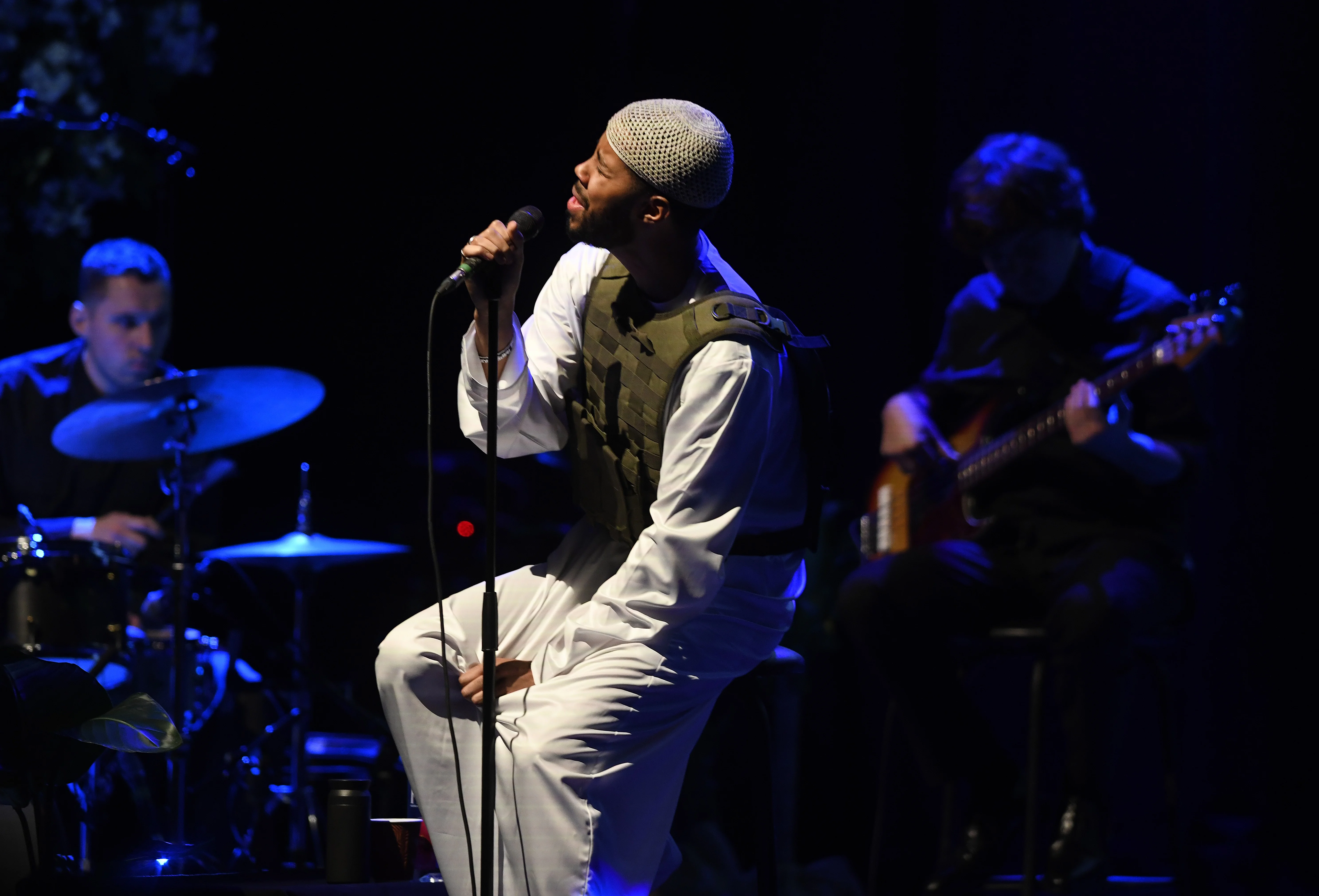 Mustafa performing live on stage in a white thobe and tactical-style vest, singing into a microphone while a drummer and bassist play behind him.