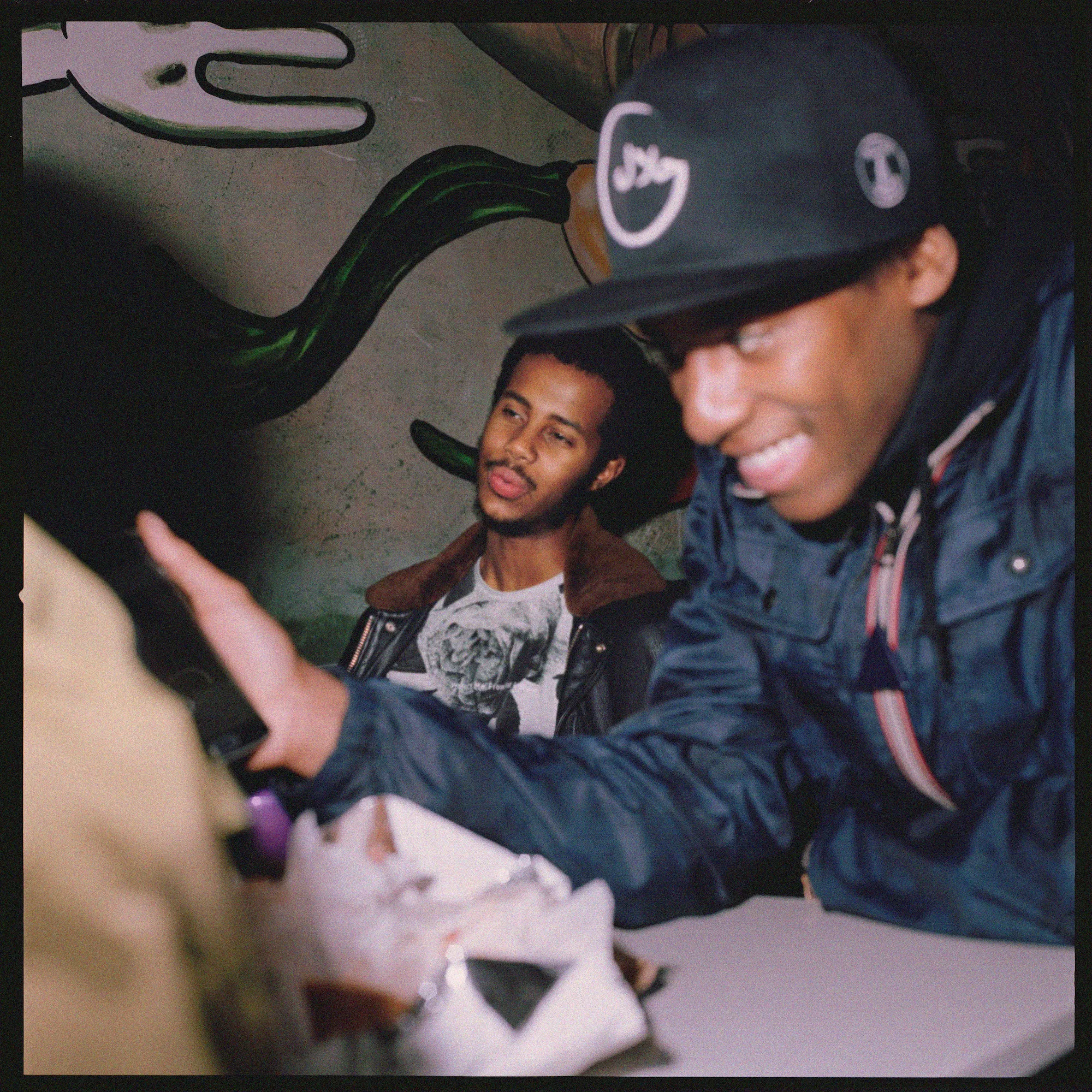 Candid photo of Mustafa sitting at a table in a leather jacket, slightly out of focus in the background while a friend in a cap smiles in the foreground.