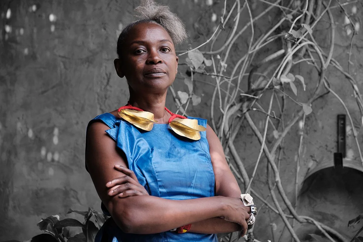 Curator Koyo Kouoh standing with folded arms in a blue dress and gold necklace, in front of a grey wall with climbing branches.