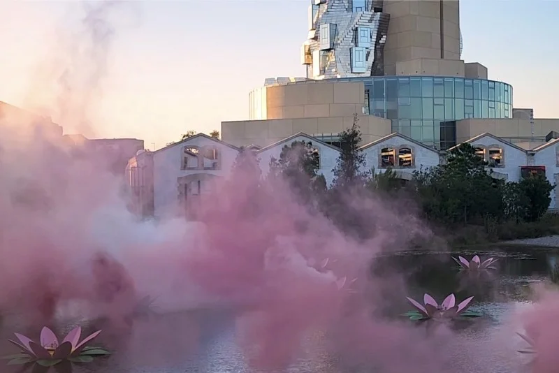 Pink smoke rising from floating lotus sculptures on a pond at sunset, with a contemporary glass and stone building in the background.