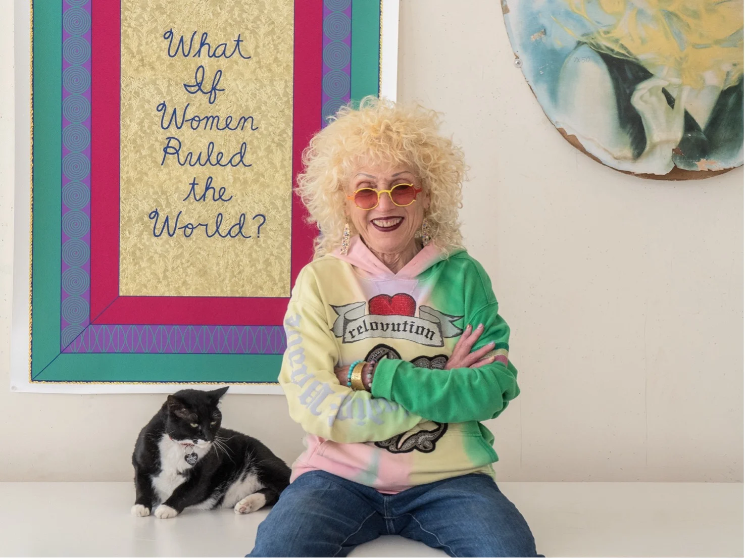 Artist Judy Chicago sits smiling with folded arms beside a black-and-white cat, in front of her work 
