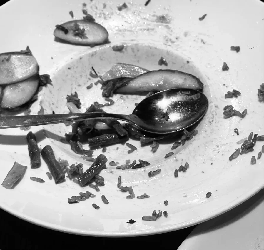 Black-and-white close-up of a nearly empty plate with leftover rice, vegetables and two spoons resting in the center.