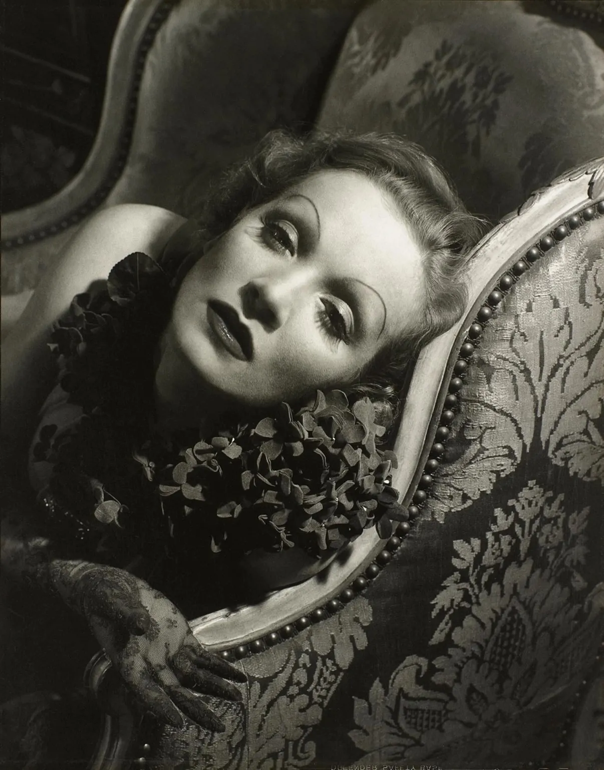 Black-and-white portrait of a woman reclining on an ornate upholstered armchair, with dramatic eye makeup, lace gloves, and a floral collar, lit in soft studio light.