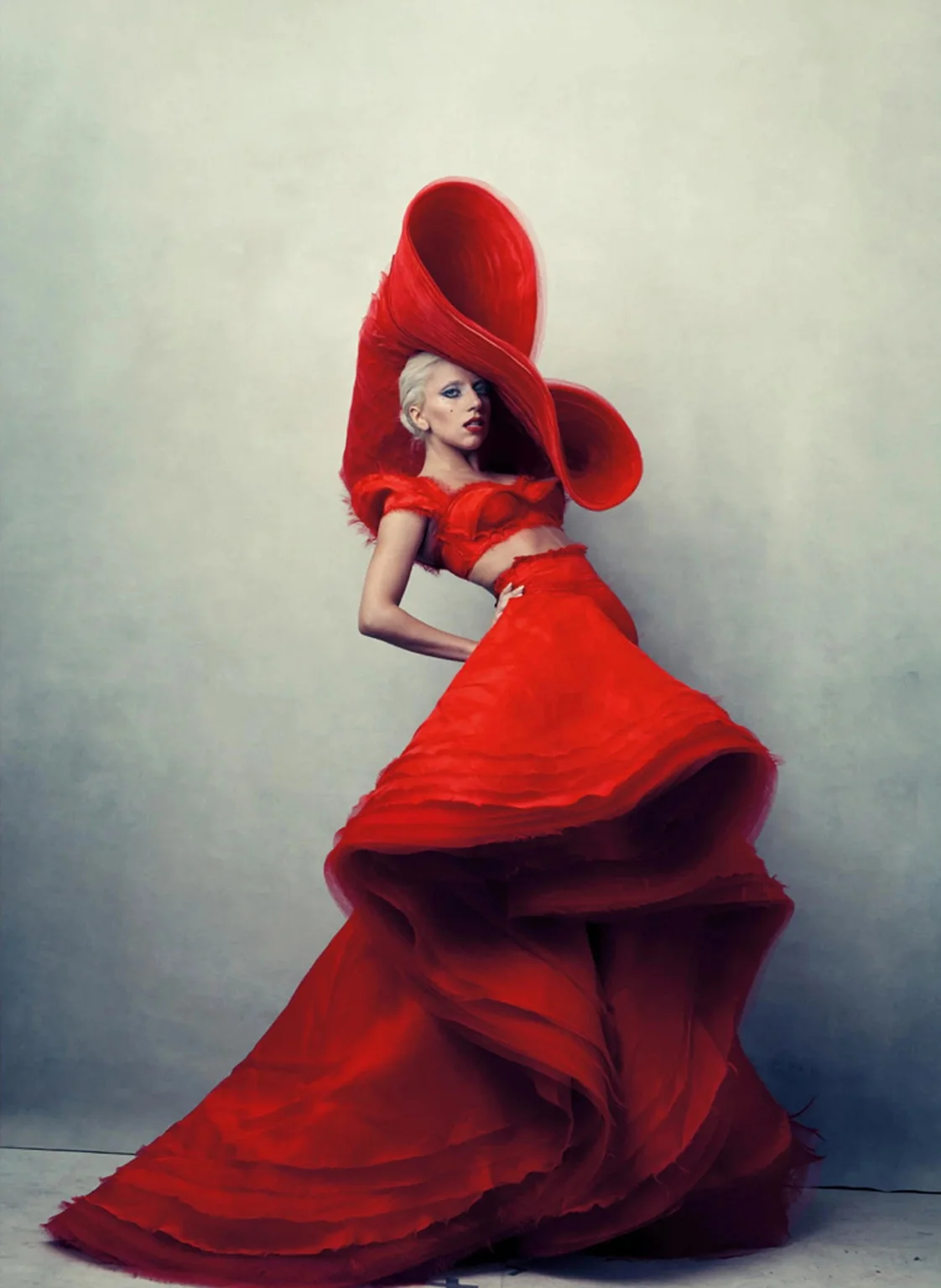 Fashion photograph of a woman in a dramatic sculptural red gown and oversized red hat against a neutral studio background.