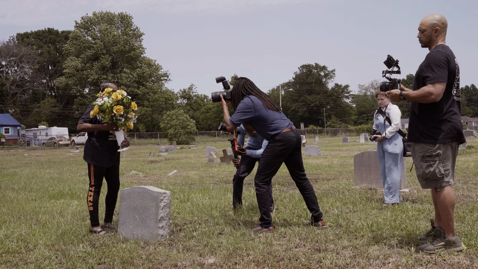 Film crew recording a mourner with yellow flowers in a Southern cemetery for Naeem Mohaiemens Through a Mirror, Darkly project.