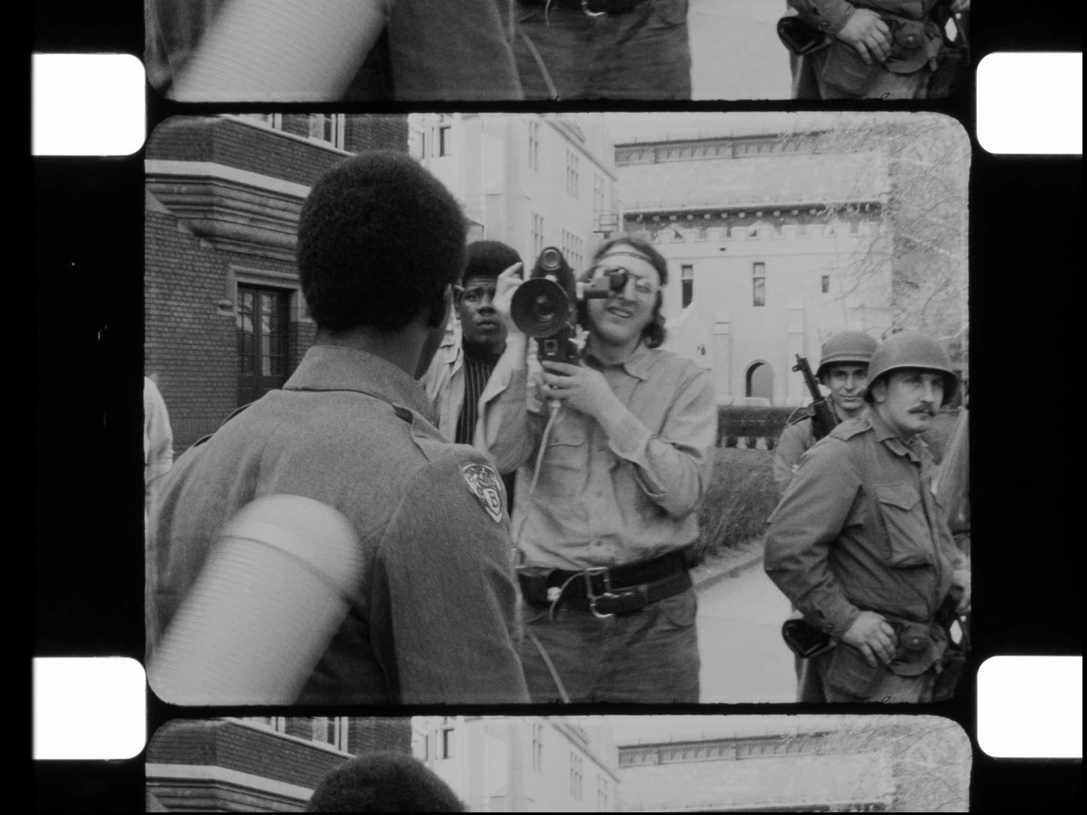 Film still from Naeem Mohaiemens Through a Mirror, Darkly, showing a Black student facing a National Guard cameraman and helmeted soldiers on a university campus.