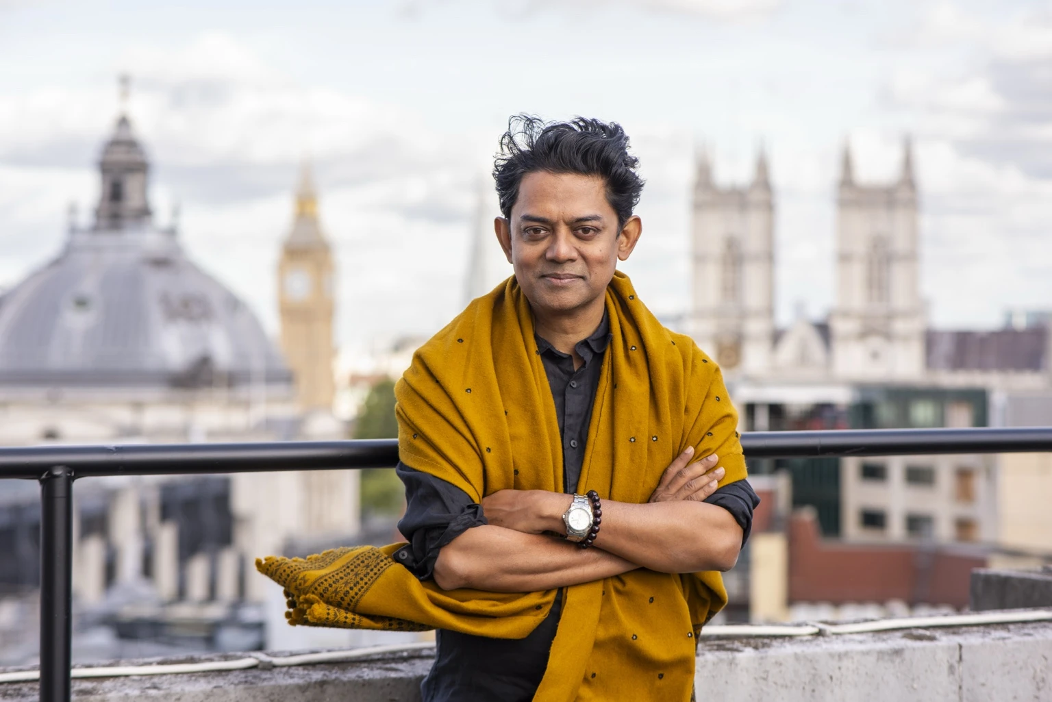Portrait of artist Naeem Mohaiemen standing on a London rooftop, wearing a mustard yellow shawl with Big Ben and Westminster Abbey softly blurred in the background.