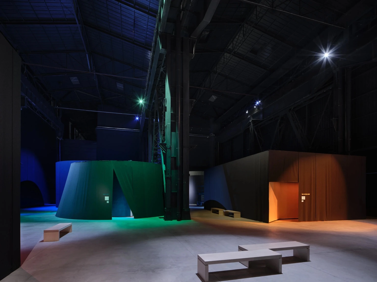 Installation view of Nan Goldins exhibition This Will Not End Well at Pirelli HangarBicocca in Milan, showing dark pavilion-like structures lit with blue, green, and orange spotlights inside a large industrial hall.