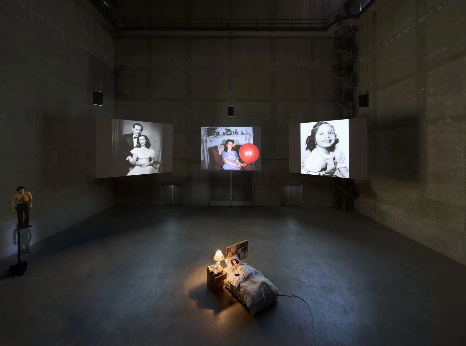 Installation view of Nan Goldins Sisters, Saints, Sibyls at Pirelli HangarBicocca in Milan, showing a single lit bed in the center of a dark hall surrounded by three large projected family photographs on the walls.