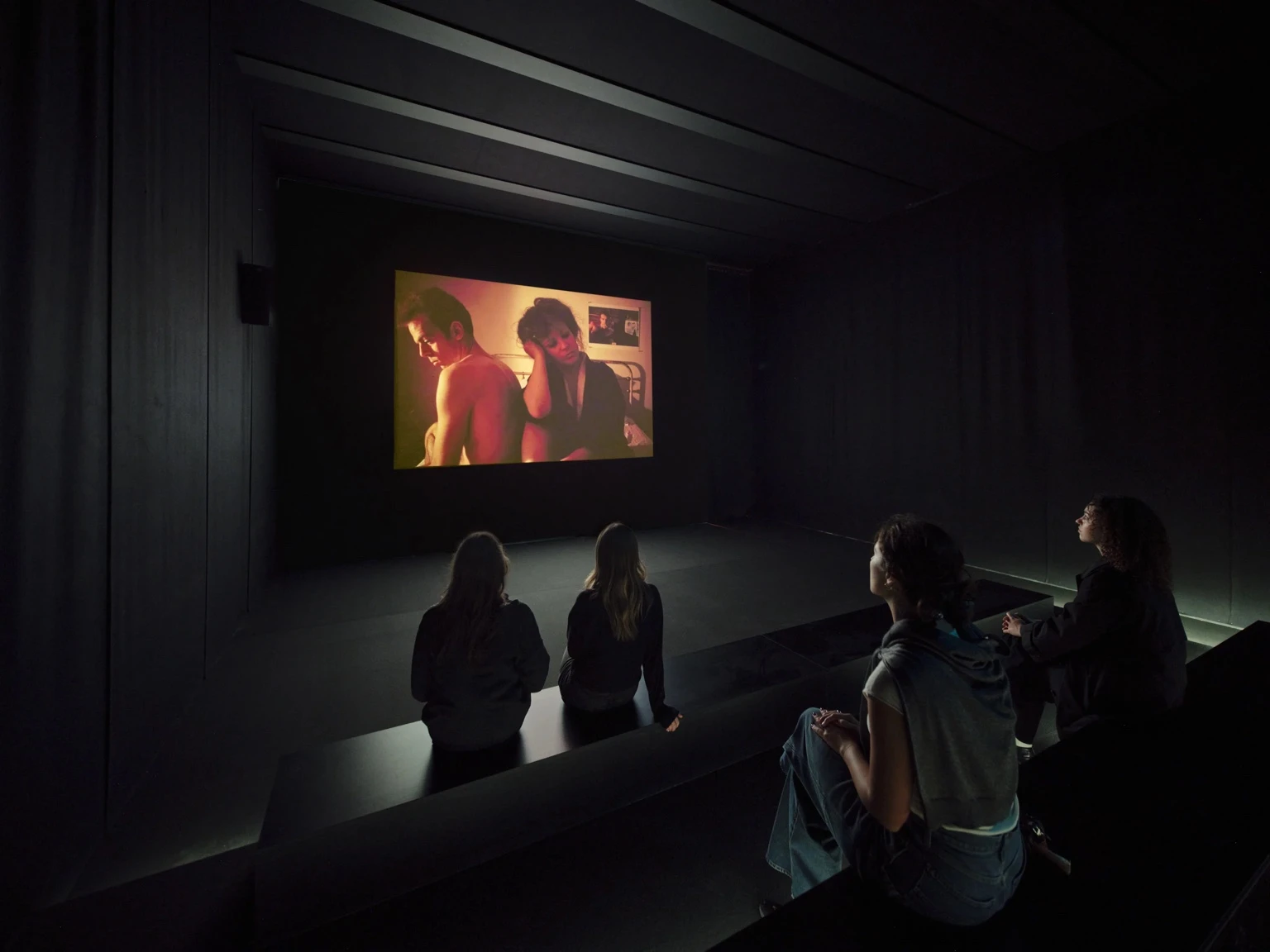 Visitors sit on dark benches in a black-box gallery watching a projected scene from Nan Goldins film The Ballad of Sexual Dependency, showing an intimate moment between two people on screen.