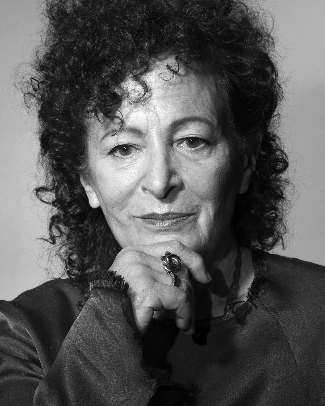 Black-and-white portrait of Nan Goldin looking at the camera, with curly hair and her hand resting near her chin, wearing a dark top and a ring