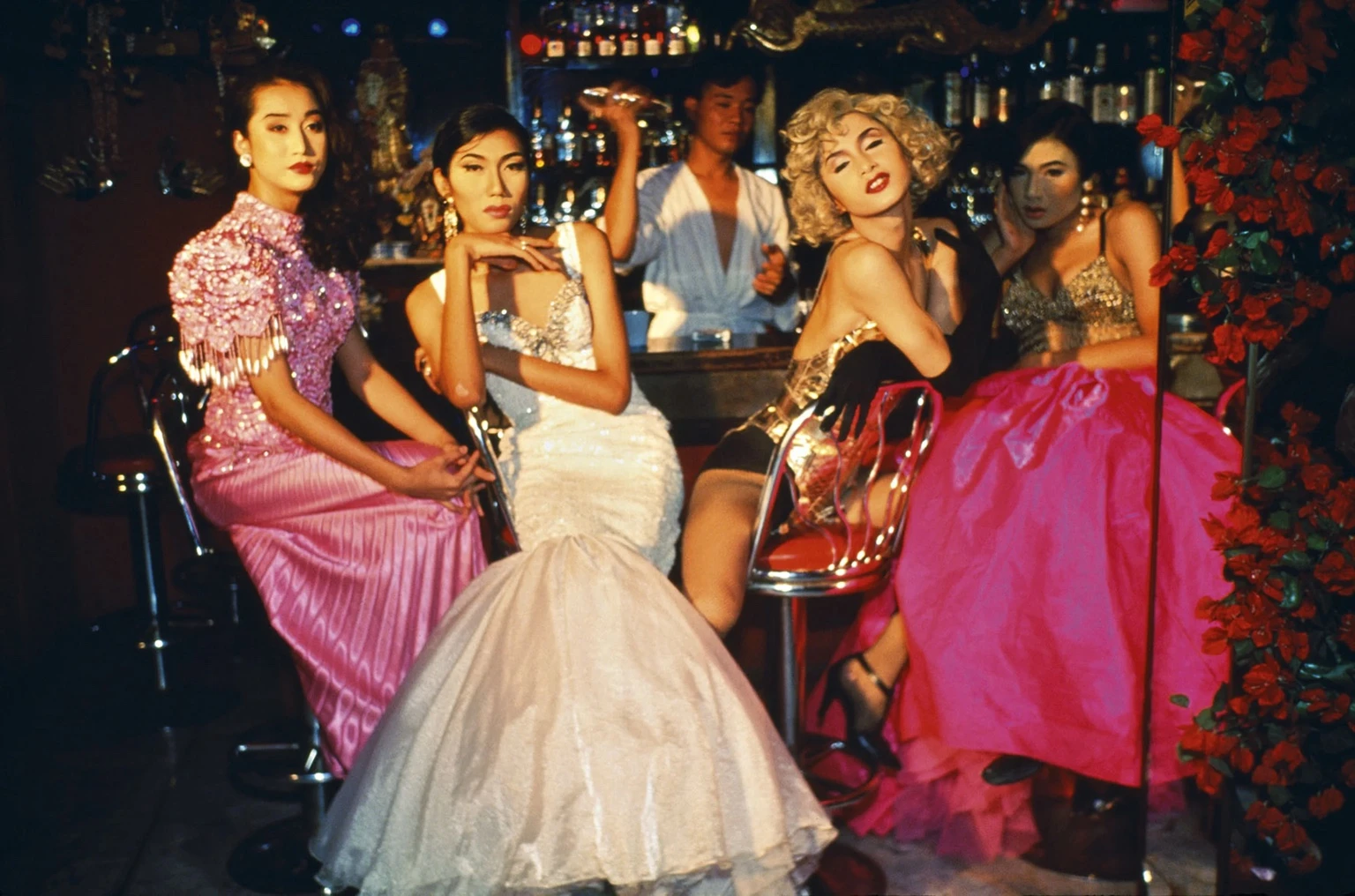 Four glamorous performers in bright gowns and evening wear pose at a bar in warm, cinematic light, in Nan Goldins photograph taken during a fashion show at Second Tip in Bangkok.