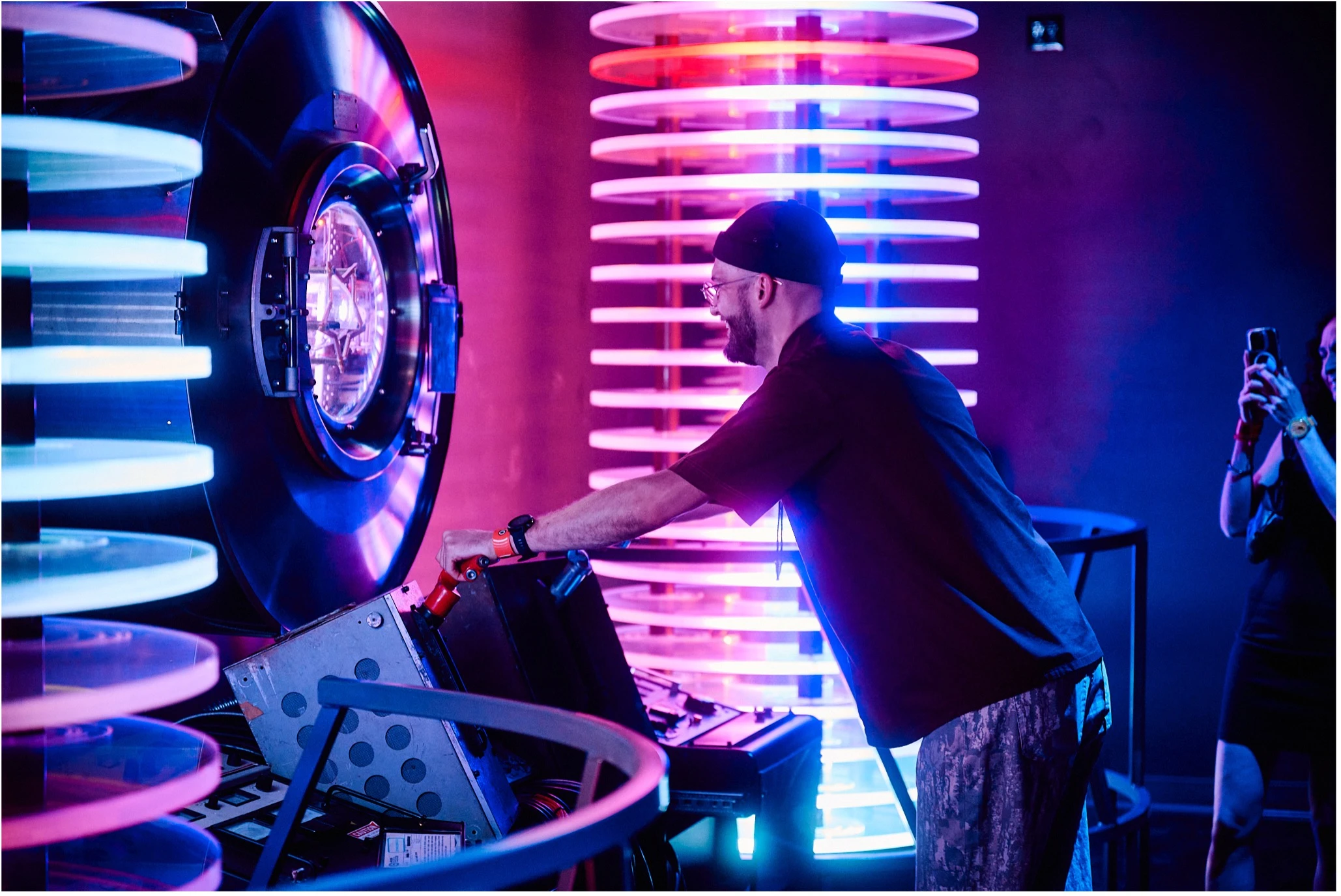 Man operating a glowing, sci-fi-style power supply interactive device surrounded by colorful neon lights while another visitor photographs the scene.