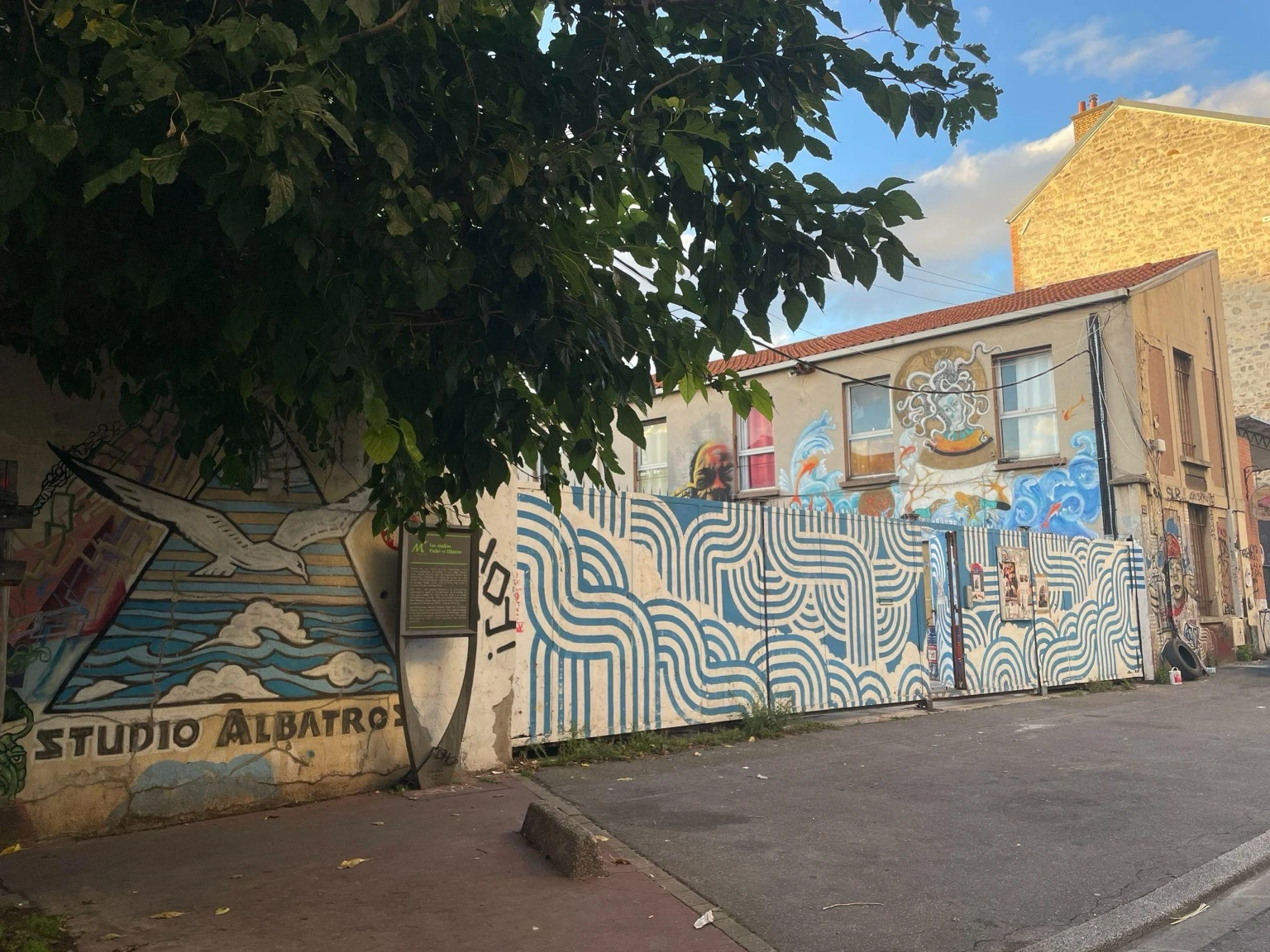 Street view of Studio Albatros in Montreuil, with colorful murals covering the walls and a blue-and-white geometric pattern painted along a fence beneath a leafy tree.