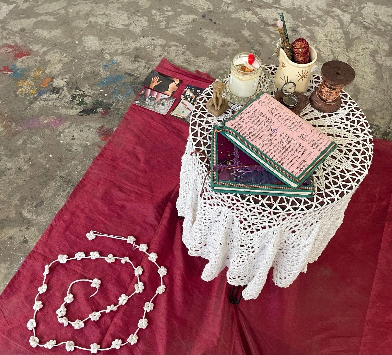 Overhead view of a small round table covered with a white crochet cloth on a red fabric, arranged with handwritten notebooks, candles, cards, and small ritual objects.