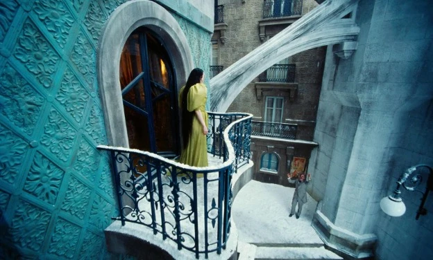 Emma Stones character stands on an ornate blue balcony overlooking a snowy courtyard while Mark Ruffalos character looks up from below in a fantastical European-style cityscape.