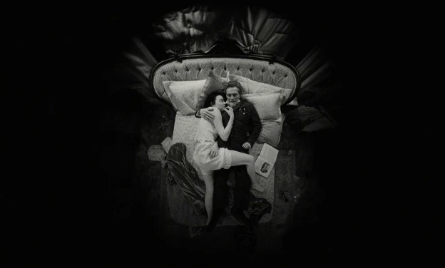 Black-and-white overhead shot of Emma Stone and Willem Dafoes characters lying side by side on an ornate bed, surrounded by pillows and blankets in a dark vignetted frame.