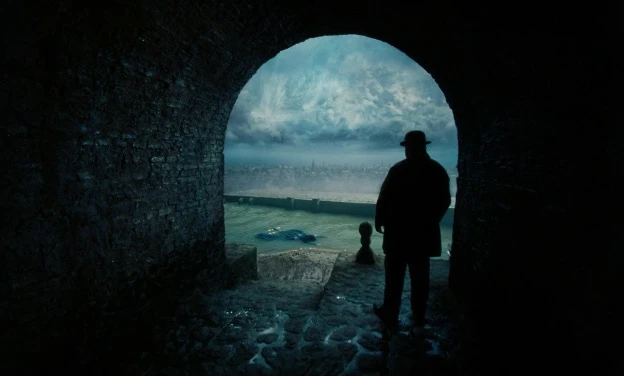 Silhouetted figure in a hat stands at the end of a dark stone tunnel, looking out over a stormy sea and distant city under a dramatic sky in Poor Things.