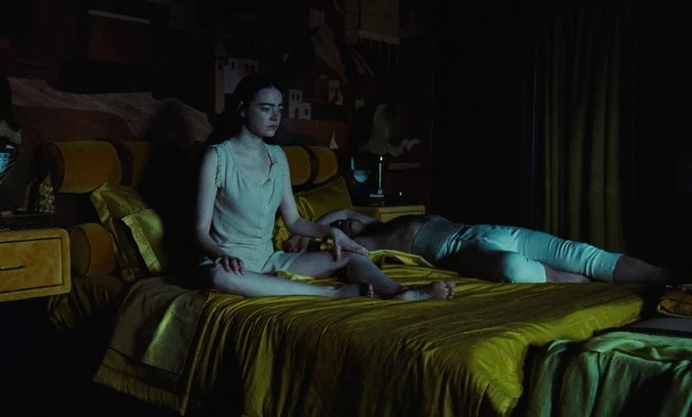 Emma Stones character sits awake on a yellow satin bed in a dimly lit room while Mark Ruffalos character lies sprawled beside her, partially out of frame.