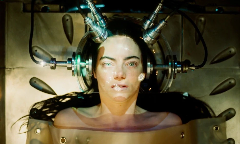 Emma Stones character lies in a futuristic medical pod with metal instruments and cables attached to a glass dome around her head, bathed in eerie golden-green light.