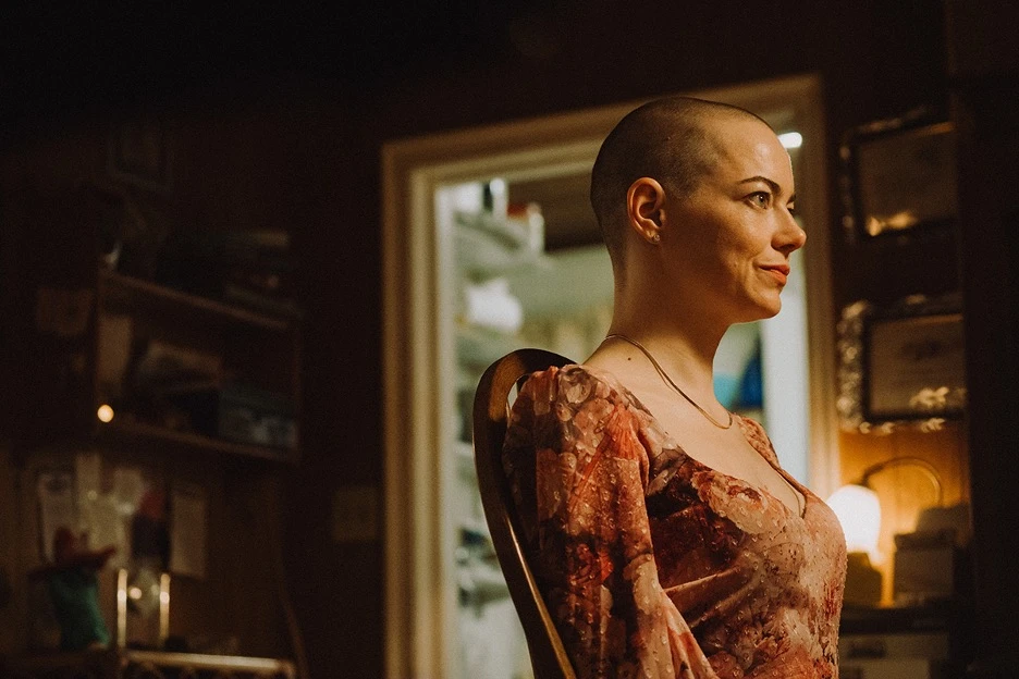 Emma Stones character with a shaved head sits in profile on a wooden chair, softly lit in a warm, cluttered interior, wearing a patterned dress.