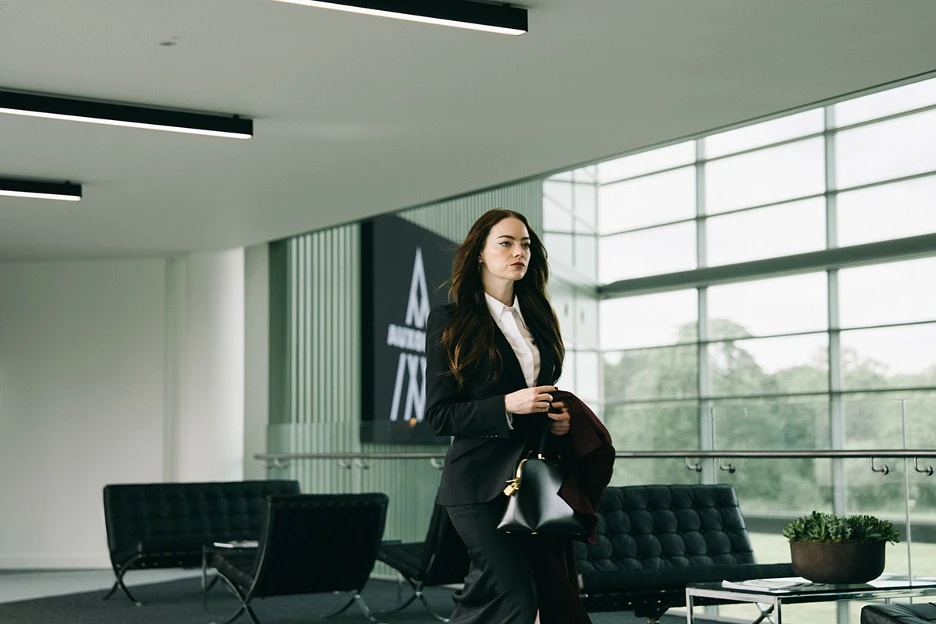 Emma Stones character in a dark business suit walks confidently through a modern glass-walled office lobby, carrying a handbag and coat.