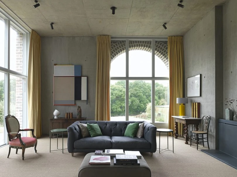 Contemporary living room with tall concrete walls, large arched window framed by mustard curtains, a dark tufted sofa with green cushions, and curated vintage furniture and art.