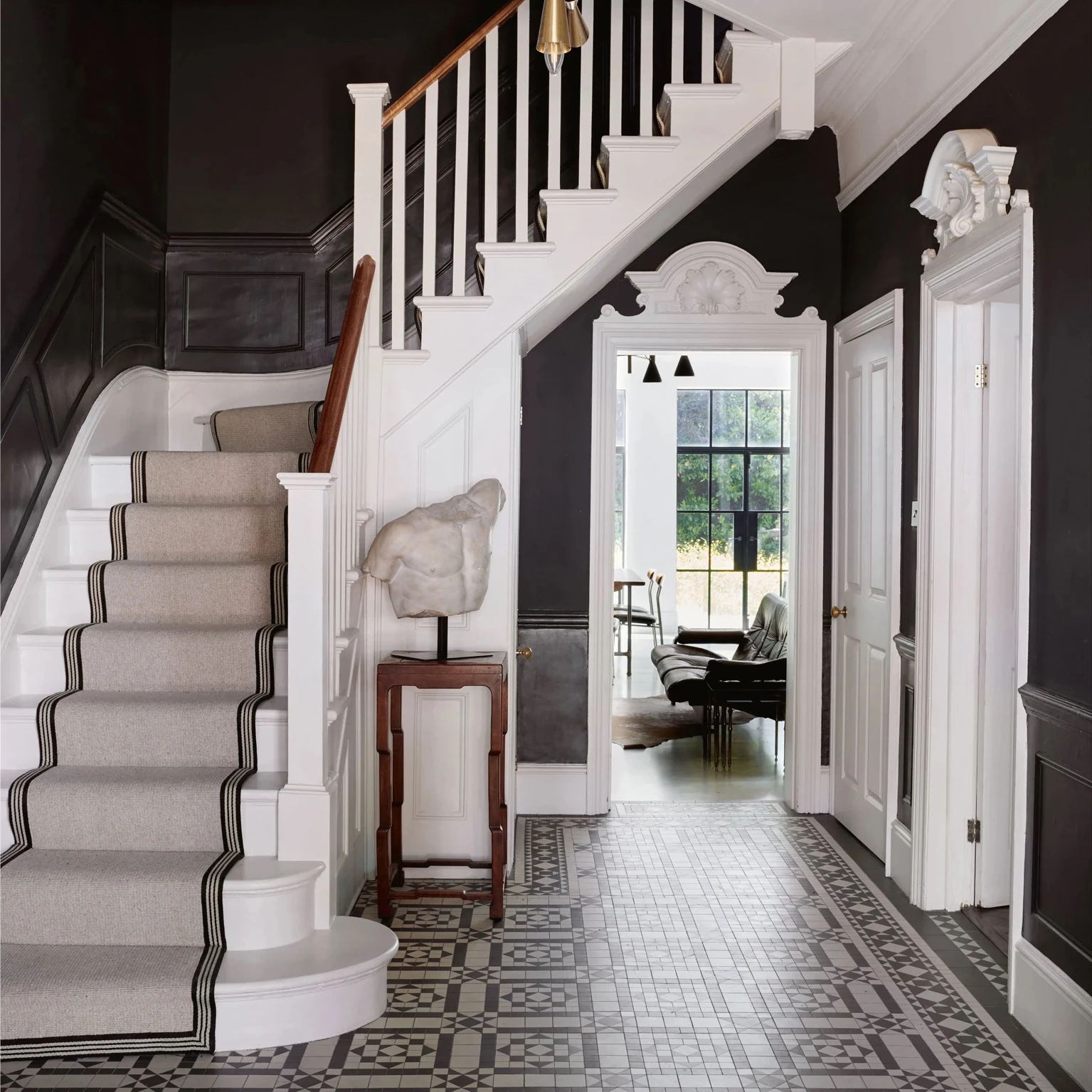 Edwardian hallway with patterned tiled floor, white staircase with a soft runner, dark painted walls, and a view through to a bright living room with large steel-framed windows.