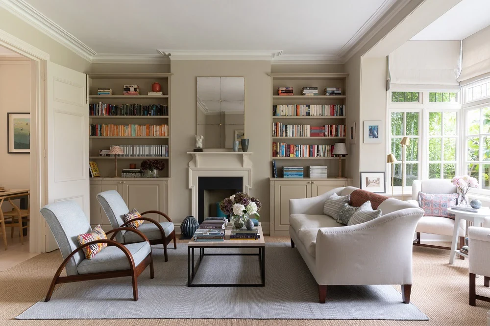 Bright, traditional living room with built-in bookshelves around a fireplace, pale upholstered sofa and armchairs, and large windows overlooking greenery.
