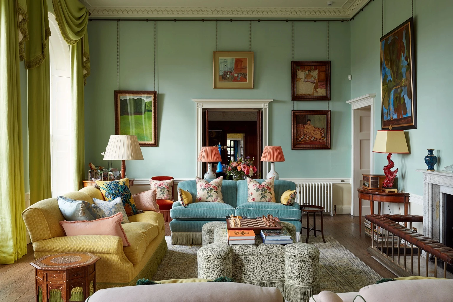 Elegant Georgian drawing room with mint-green walls, colourful sofas, floral cushions, hanging paintings, and classic furniture arranged around a central coffee table.