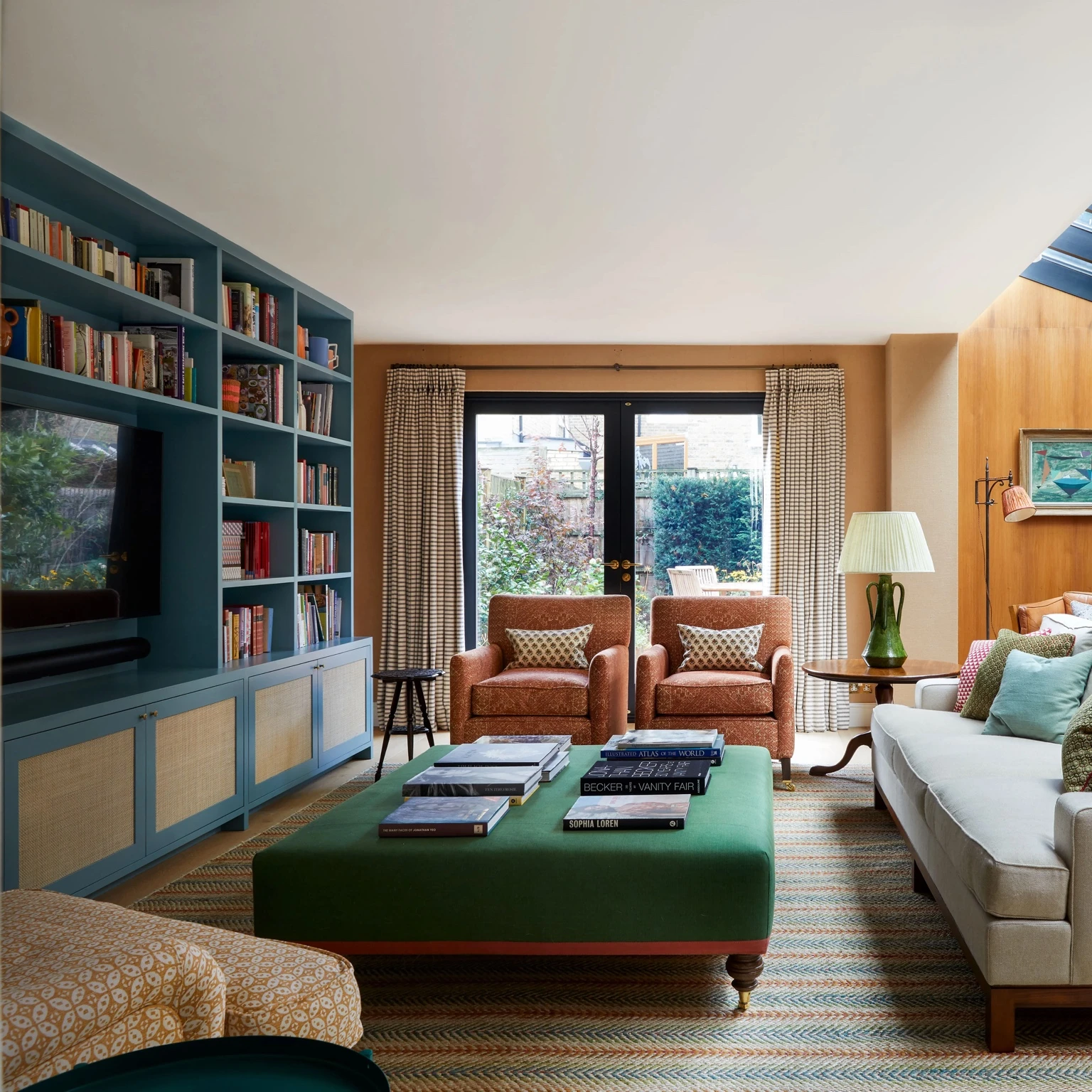 Warm, book-lined living room by Brandon Schubert, with teal built-in shelves, terracotta armchairs, a large green ottoman, and soft layered textiles.