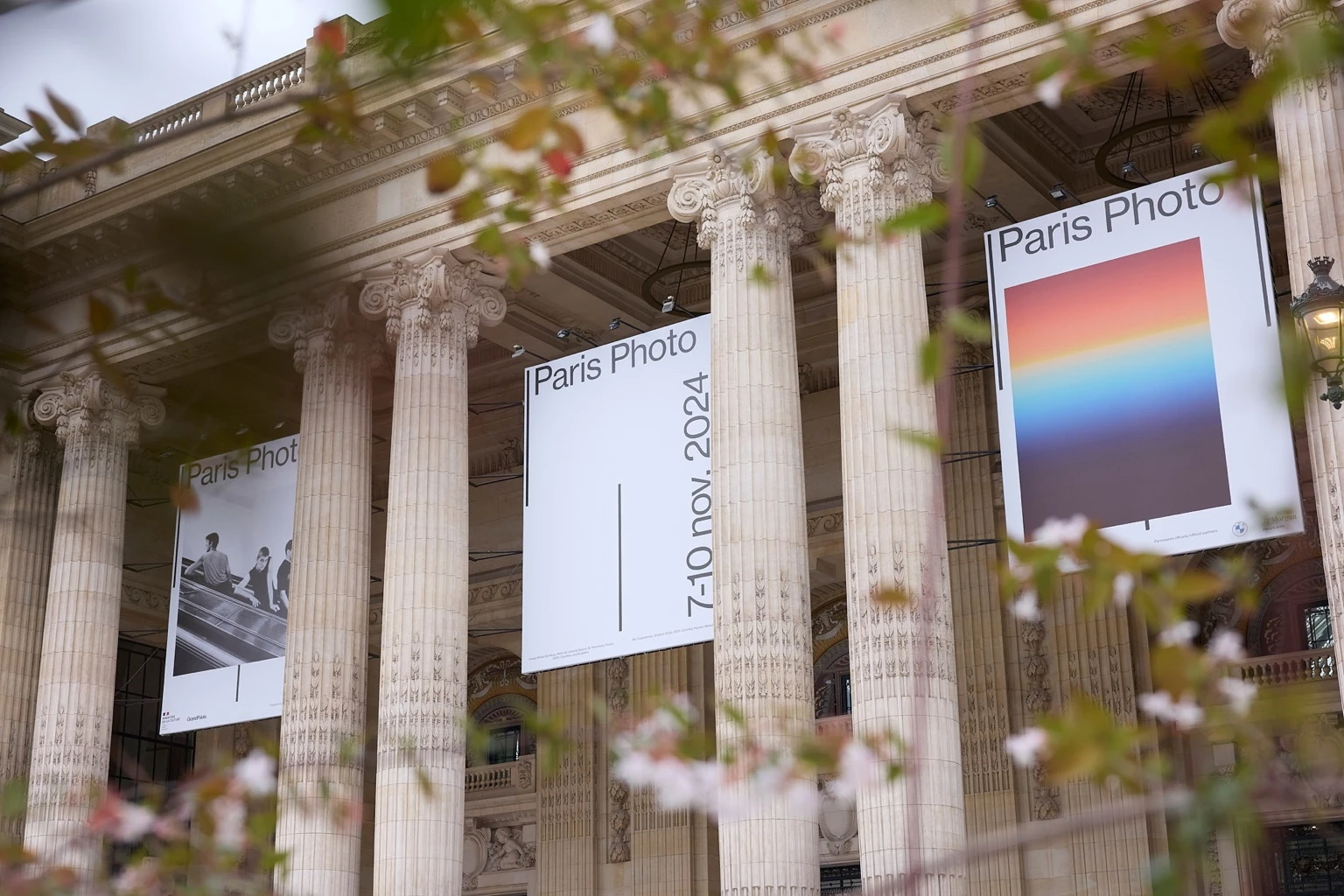 Large Paris Photo 2024 banners hanging between the classical stone columns of a historic Parisian building, seen through slightly blurred branches with leaves and flowers in the foreground.