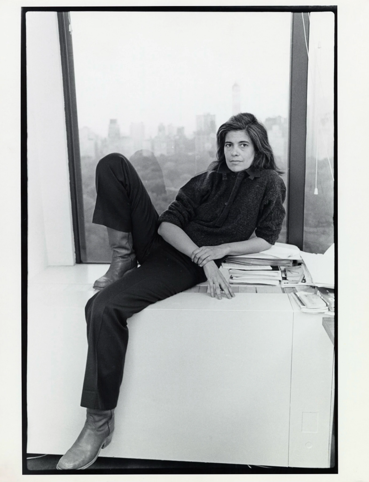 Black-and-white portrait of Susan Sontag reclining on a desk beside a stack of papers, photographed inside her New York apartment with the city skyline visible through a window.