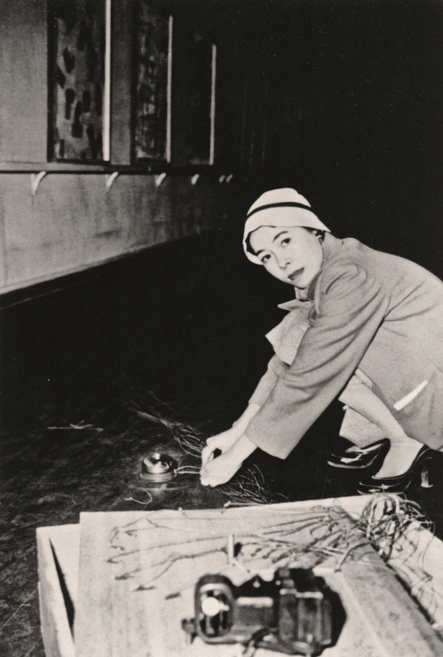 Black-and-white photograph of Atsuko Tanaka installing her sound work Bell, shown crouching on the floor connecting wires to a small bell as part of a participatory installation.