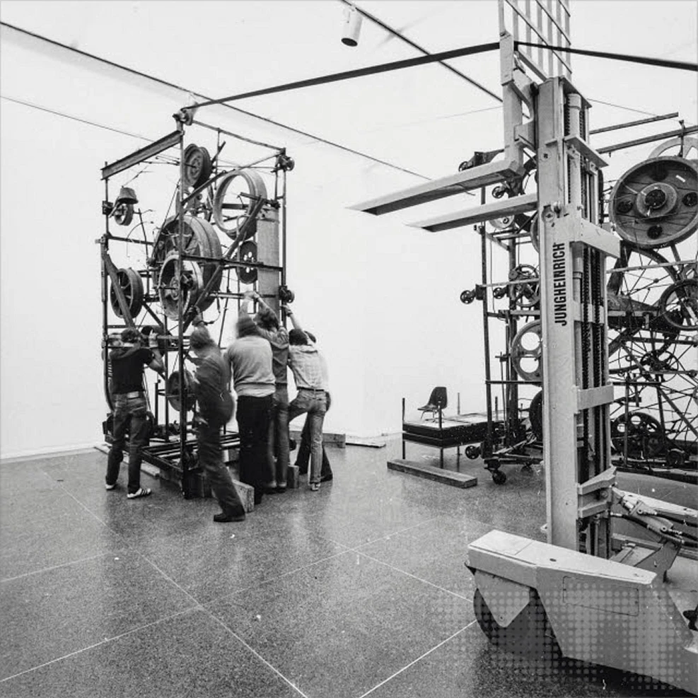 Black-and-white installation photograph of Jean Tinguely’s kinetic sculpture Mta-Harmonie II, showing a large mechanical structure of wheels and metal components assembled in a gallery space.