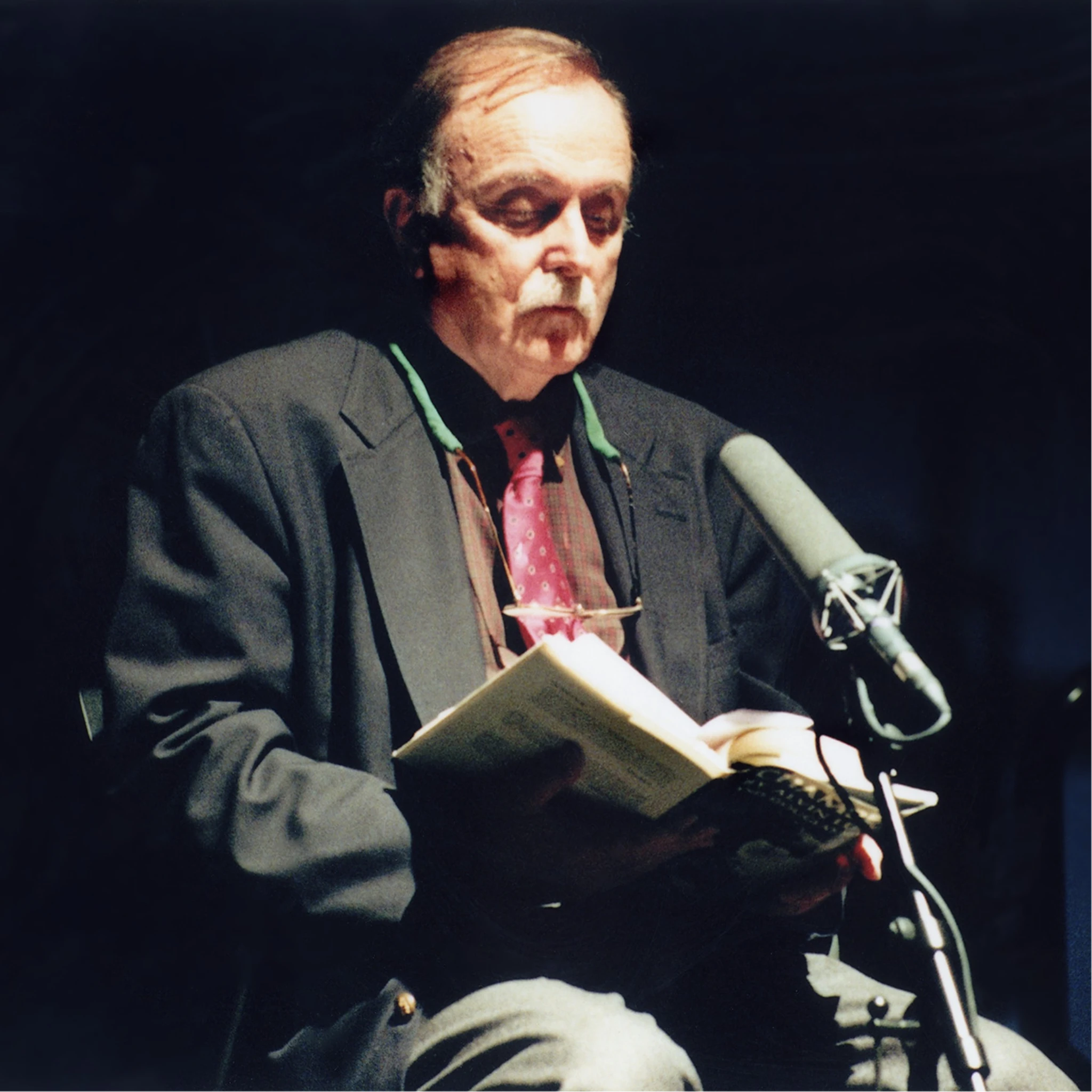 Portrait of Alvin Lucier reading from a book into a microphone during a performance of I Am Sitting in a Room, emphasizing voice, repetition, and acoustic space.