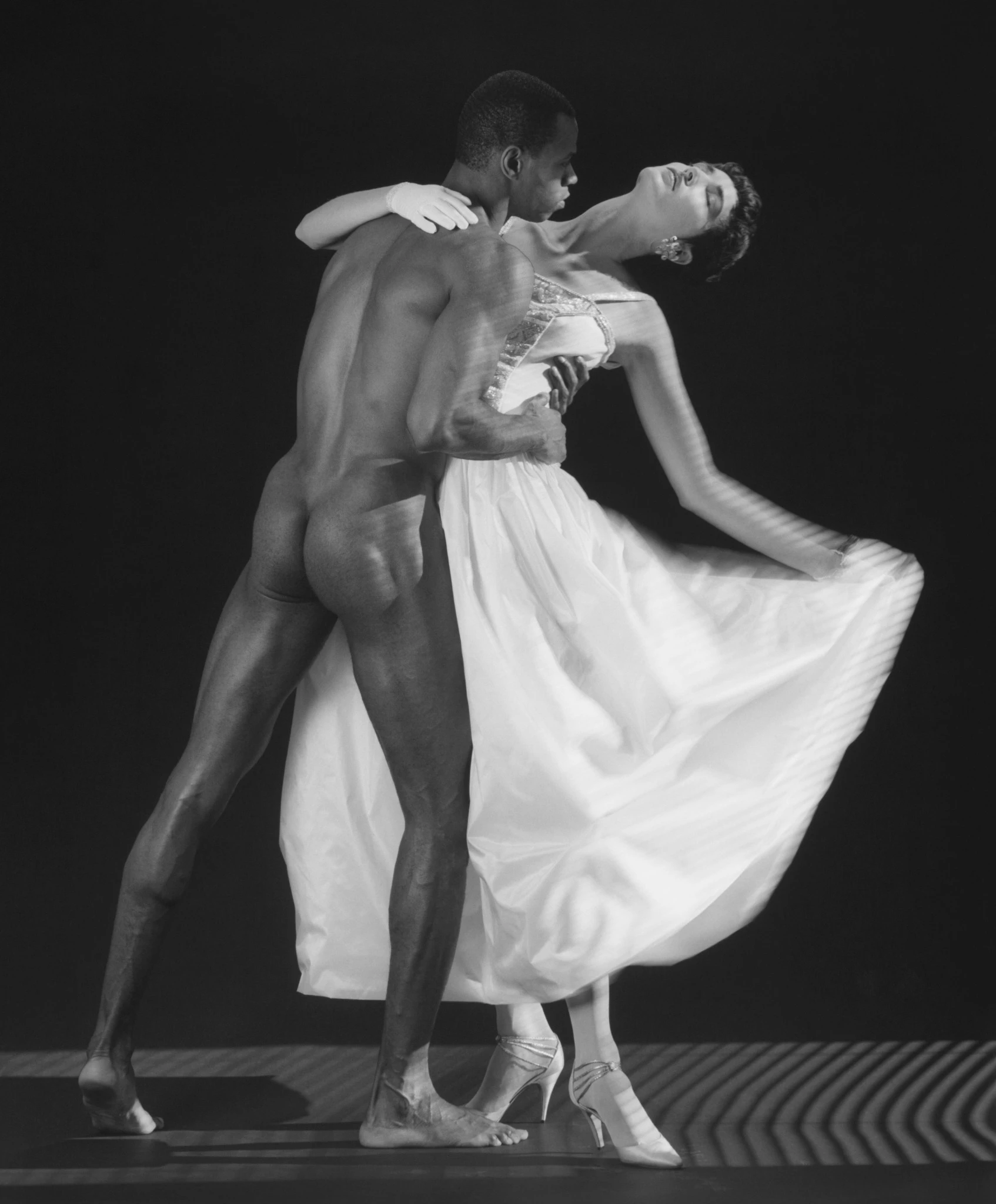 Black and white studio photograph of a nude male dancer holding a woman in a flowing white dress as she arches backward in an elegant pose against a dark background. Robert Mapplethorpe, Thomas and Dovanna, 1986 Robert Mapplethorpe Foundation