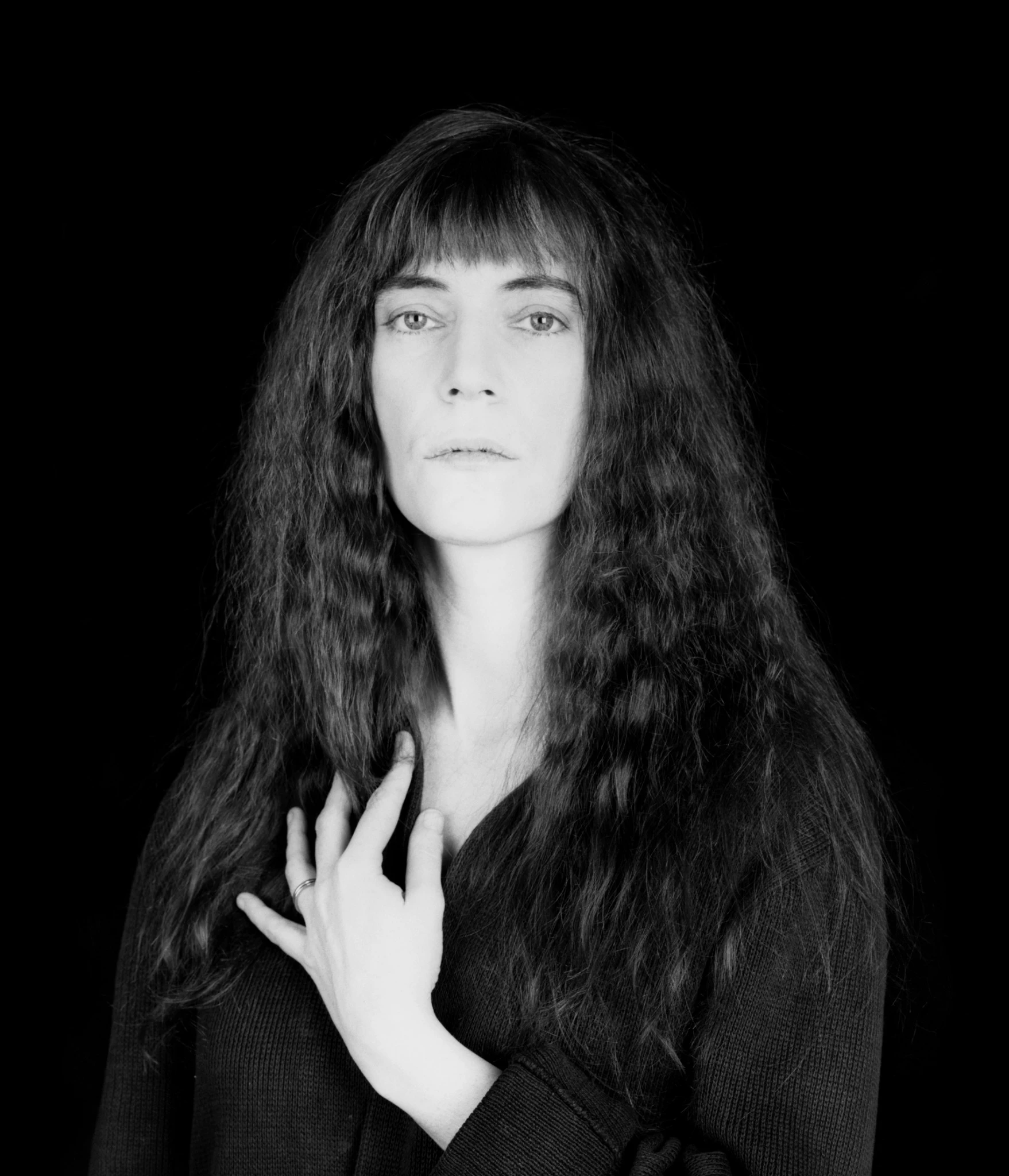 Black and white portrait of Patti Smith with long textured hair and straight bangs, looking directly at the camera against a dark background with one hand resting near her collarbone. Robert Mapplethorpe, Patti Smith, 1986 Robert Mapplethorpe Foundation