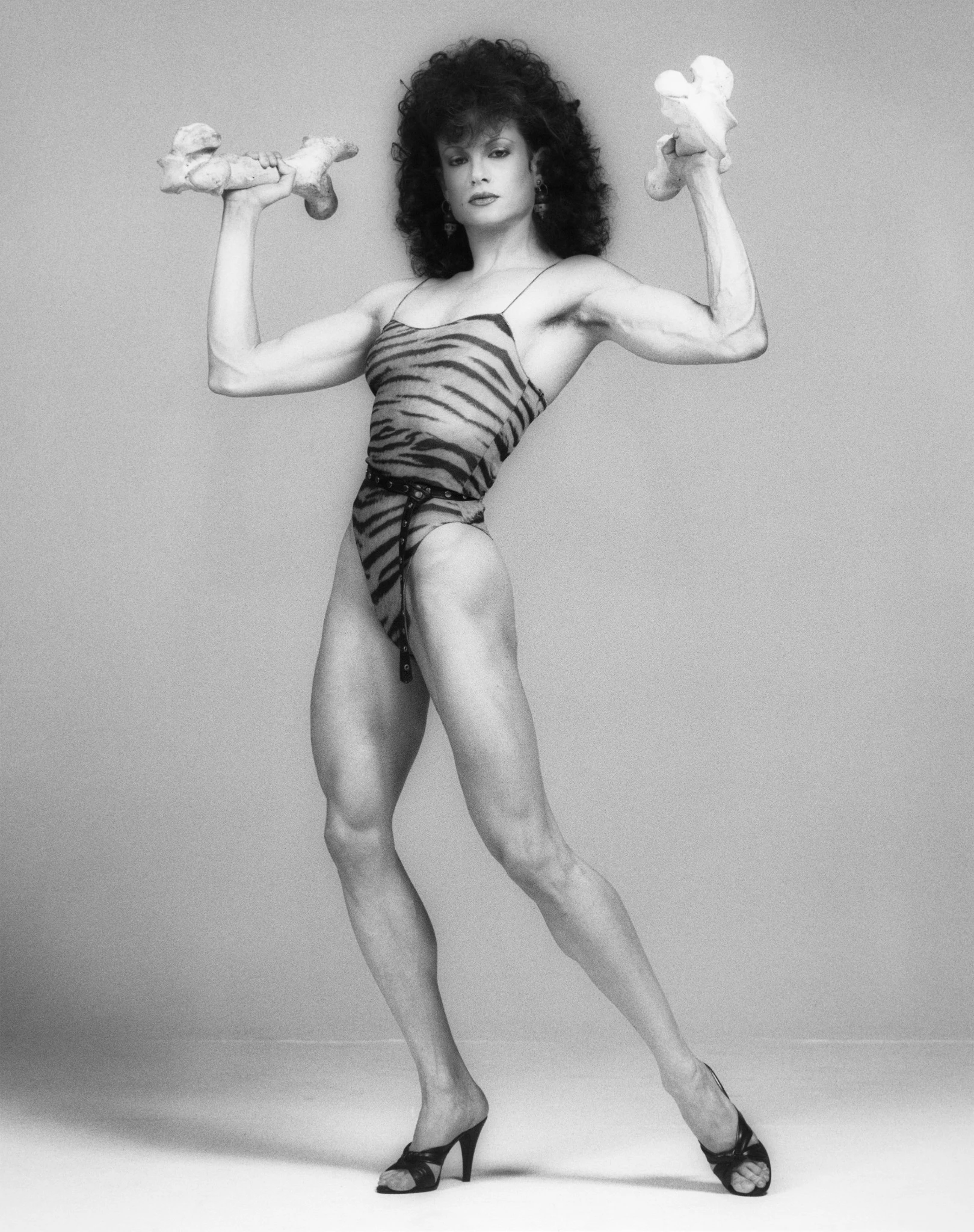 Black and white studio portrait of bodybuilder Lisa Lyon posing confidently while holding two bones like weights, wearing a patterned bodysuit and heels against a plain background. Robert Mapplethorpe, Lisa Lyon, 1983 Robert Mapplethorpe Foundation