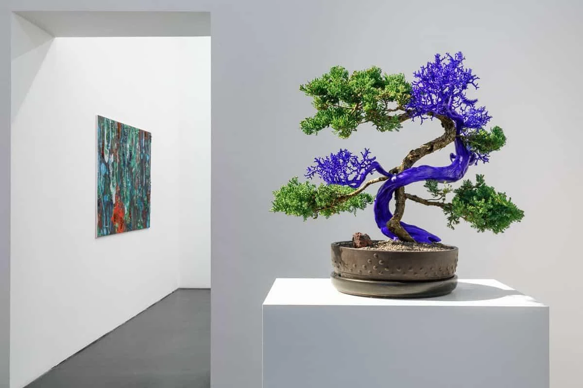 Installation view of Variantology by Agnieszka Kurant, featuring a bonsai-like sculptural tree with vivid purple sections displayed on a white pedestal in a gallery space, with a colorful abstract painting visible on the adjacent wall.