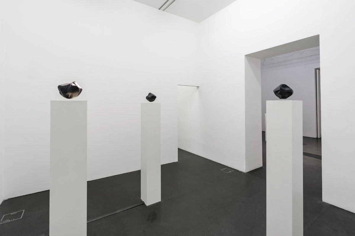 Installation view of Variantology by Agnieszka Kurant showing three dark sculptural forms displayed on tall white pedestals in a minimalist gallery space, with a doorway leading to another room in the background.