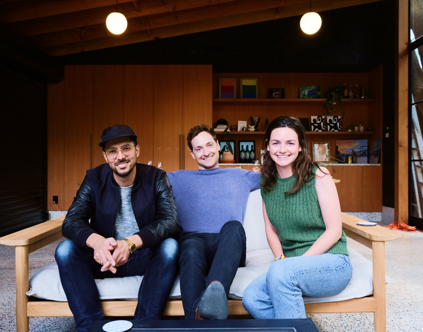 Three Peggy team members smiling together seated on wooden couch in modern studio space