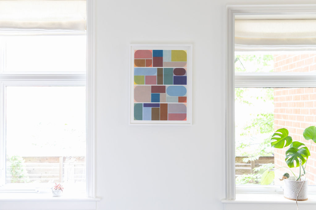 Colorful abstract geometric shapes print in white frame hung between two windows with monstera plant